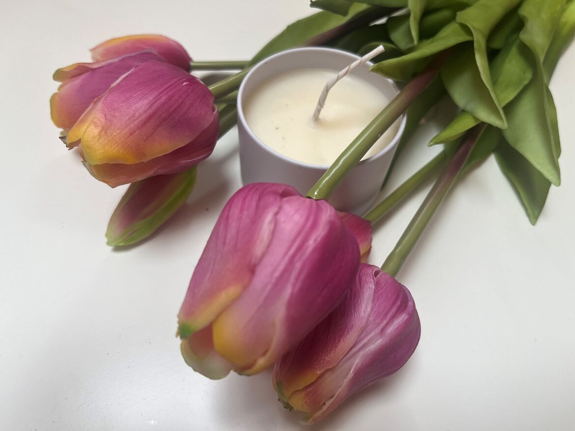 Vier violett-gelbe Tulpen mit grünen Blättern liegen neben einer weißen DIY-Massagekerze auf einer glatten, weißen Oberfläche. | © BILDderFRAU/Madeleine Liedke 