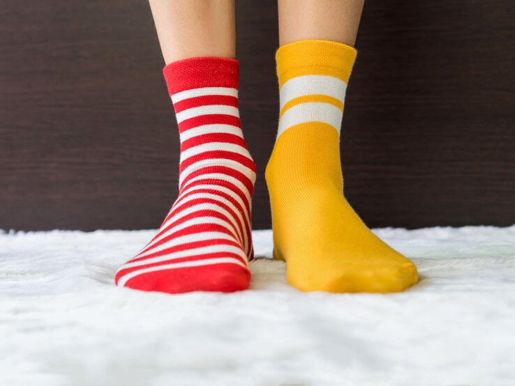 Zwei Füße in ungleichen Socken, eine rot-weiß gestreift, die andere gelb mit weißen Streifen, auf flauschigem Teppich vor dunklem Hintergrund.