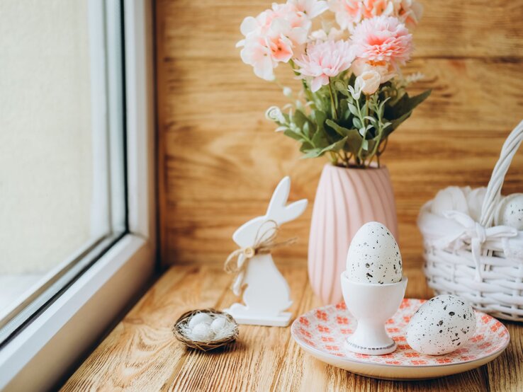 Frühlingshaft gedeckte Fensterbank mit Ostereiern, einem kleinen Nest, pastellfarbenen Blumen und einem weißen Korb vor Holzoptik.