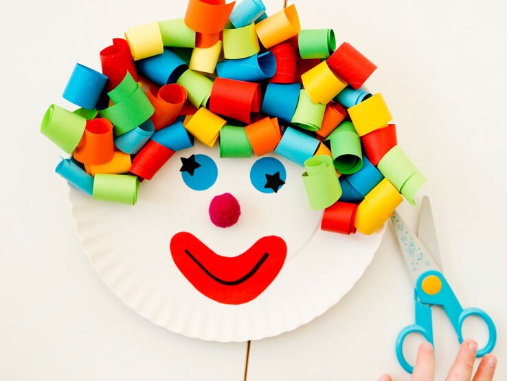 Buntes Clown-Gesicht aus einem Pappteller mit Locken aus gerolltem Papier, roten Lippen und Pompon-Nase, daneben eine Schere und eine Kinderhand.