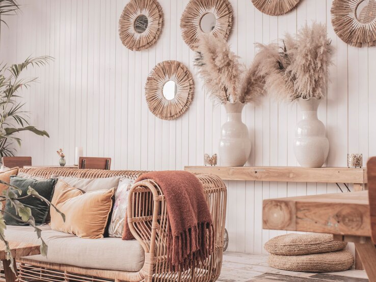 Gemütliches Boho-Wohnzimmer mit Rattansofa, warmen Kissen, Pampasgras in Vasen, Bastspiegeln an weißer Wand und rustikalen Holzmöbeln.