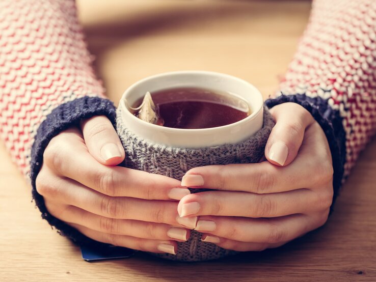 Zwei Hände mit zarten, lackierten Fingernägeln umschließen eine Teetasse mit Teebeutel darin mit gestricktem Überzug auf einem Holztisch.