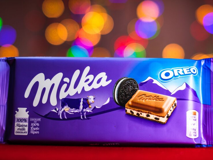 Eine Tafel Milka-Oreo-Schokolade in lilafarbener Verpackung mit Alpenmotiv und Milka-Kuh, vor buntem, unscharfem Lichterhintergrund.