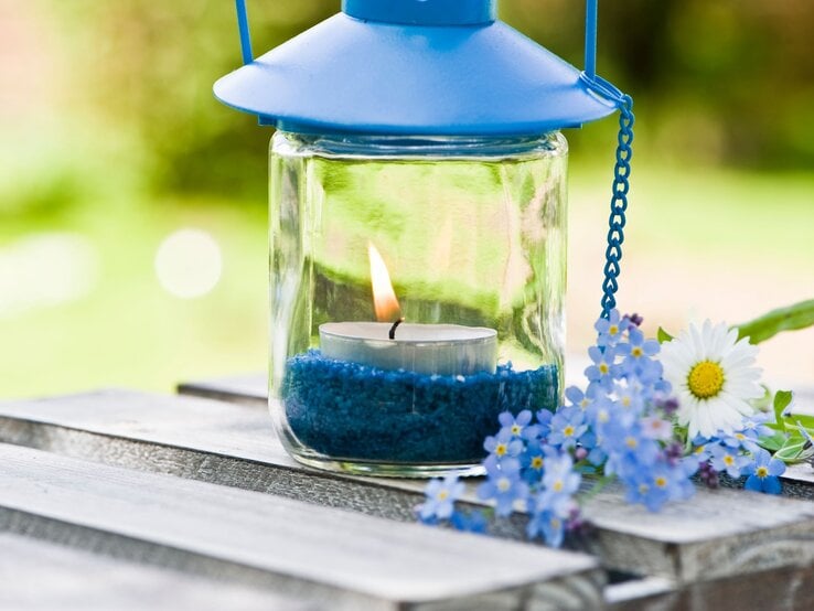 Ein Windlicht mit blauer Metallhaube und brennender Kerze steht auf einem Holztisch, umgeben von blauen Blüten.