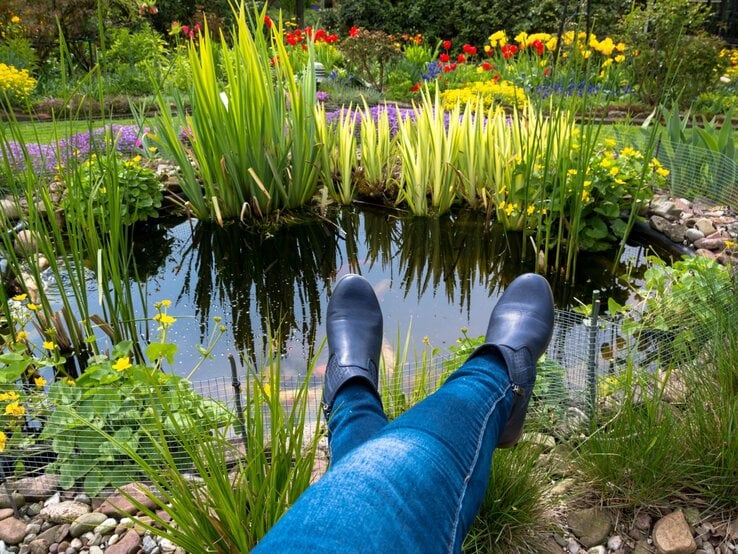 Eine Person in blauen Jeans und schwarzen Stiefeletten streckt entspannt die Beine aus und genießt den Blick auf einen idyllischen Gartenteich.