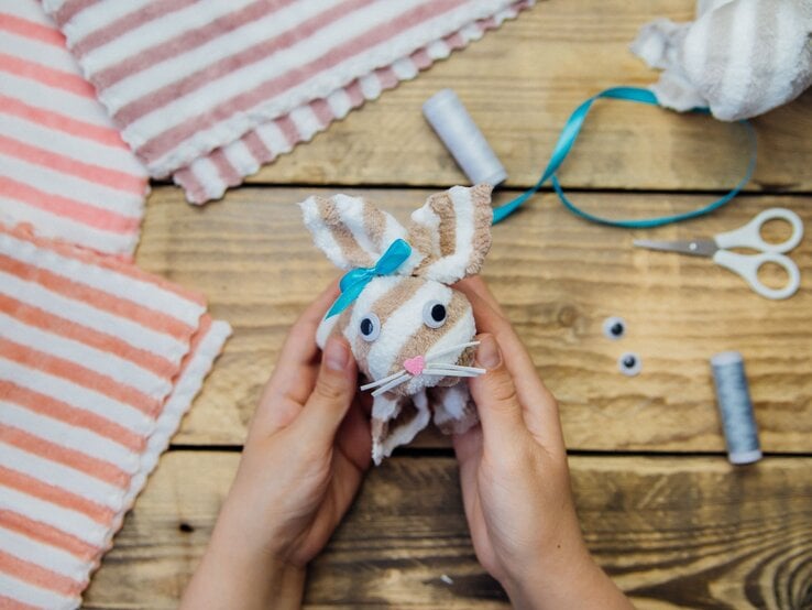 Zwei Hände halten ein selbstgebasteltes Stoffhasen zu Ostern aus gestreiftem Handtuch mit blauer Schleife auf Holztisch.