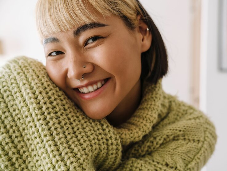 Fröhliche Frau mit kurzen, zweifarbigen Haaren und Piercings, die einen grünen Strickpullover trägt.