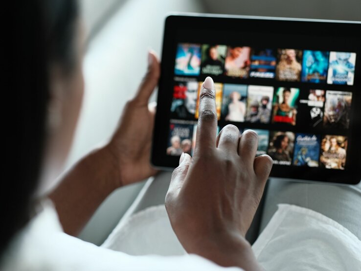 Eine Person mit dunkler Hautfarbe wählt auf einem Tablet im Querformat einen Film aus einer Streaming-Bibliothek aus.
