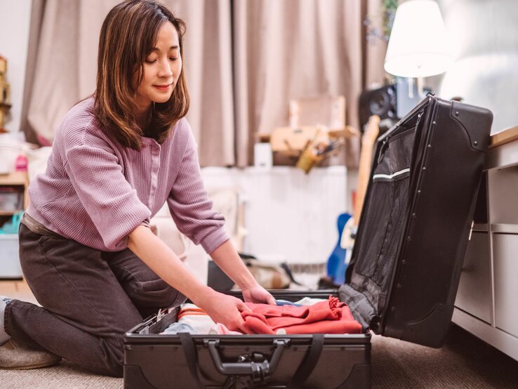 Lächelnde Frau in lässiger Kleidung kniet in einem gemütlichen, hell erleuchteten Zimmer vor einem geöffneten Koffer, um ihn zu packen.