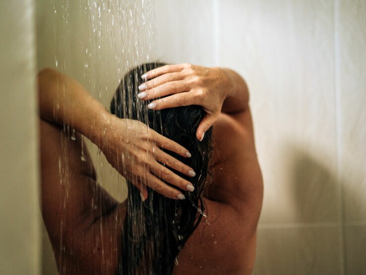 Eine Frau mit nassen Haaren steht in der Dusche und hält sich die Hände über den Kopf, während Wasser auf sie herabströmt.