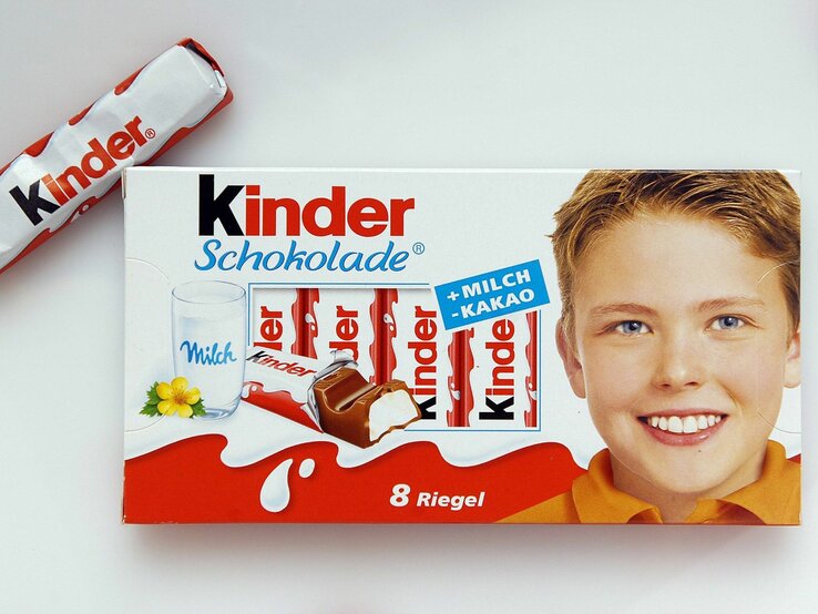 Packung Kinder Schokolade. Auf der linken Seite der Verpackung ist ein Glas Milch mit einem gelben Blümchen abgebildet, daneben sind einige Schokoladenriegel zu sehen. Ein einzelner Schokoladenriegel ist neben der Verpackung platziert. Auf der rechten Seite der Verpackung ist das lächelnde Gesicht eines Jungen zu sehen. 