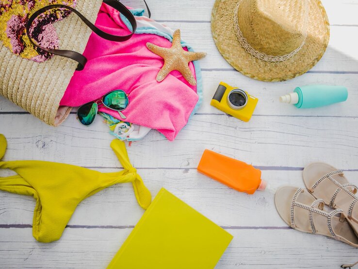 Eine gelbe Bikinihose, Sonnencreme, Kamera und Sonnenhut liegen auf einem Holztisch neben einer Strandtasche aus Stroh mit Handtuch und Sonnenbrille.