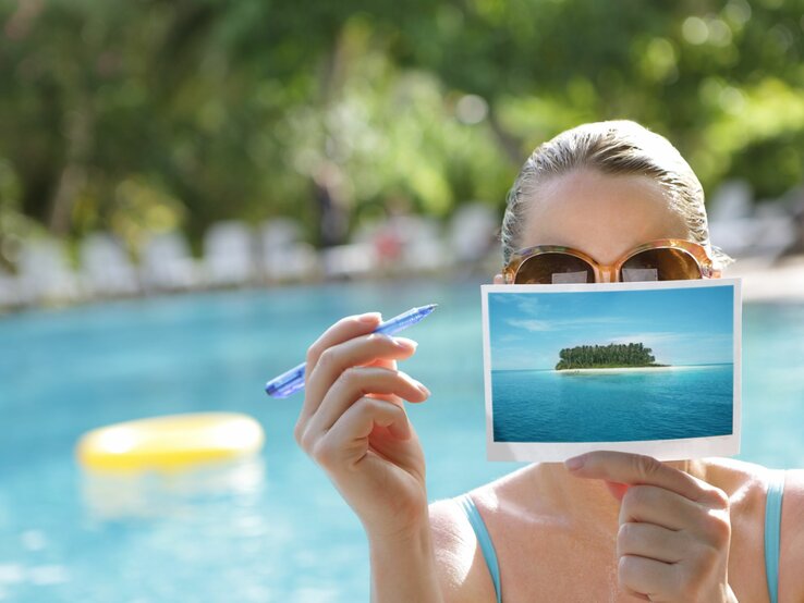 Eine Frau mit brauner Sonnenbrille hält eine Postkarte von einer Insel und einen blauen Stift vor einem Pool.
