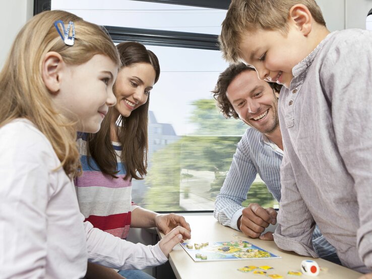 Eine lachende Familie spielt ein Brettspiel am Tisch eines fahrenden Zuges, im Hintergrund sind Bäume.