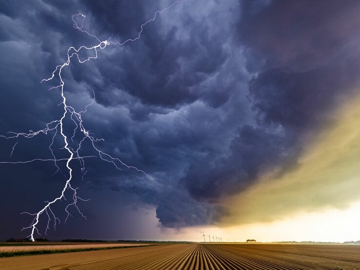 Ein Blitz erhellt dramatisch den dunklen Himmel über einem abgeernteten Feld mit Windrädern im Hintergrund.