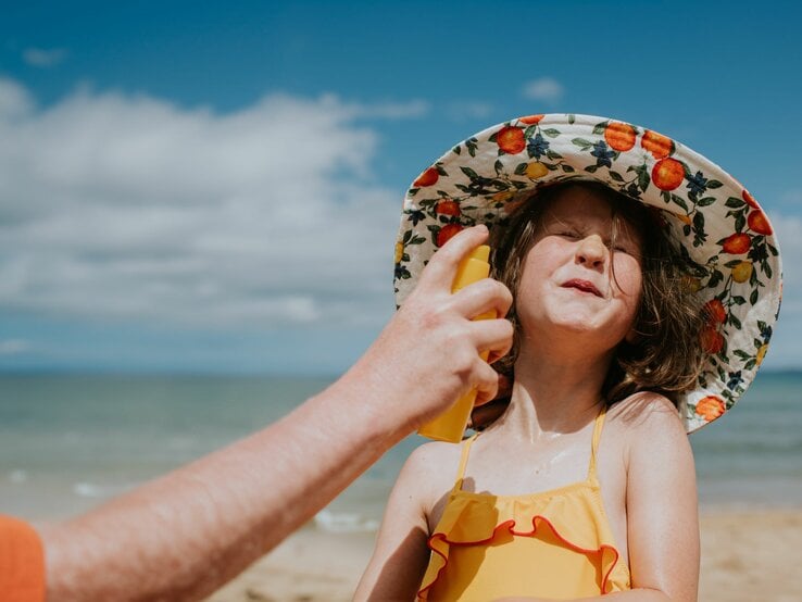 Ein kleines Mädchen in einem gelben Badeanzug und Hut mit Fruchtmuster kneift lachend die Augen zu, während ihr jemand Sonnencreme ins Gesicht sprüht.