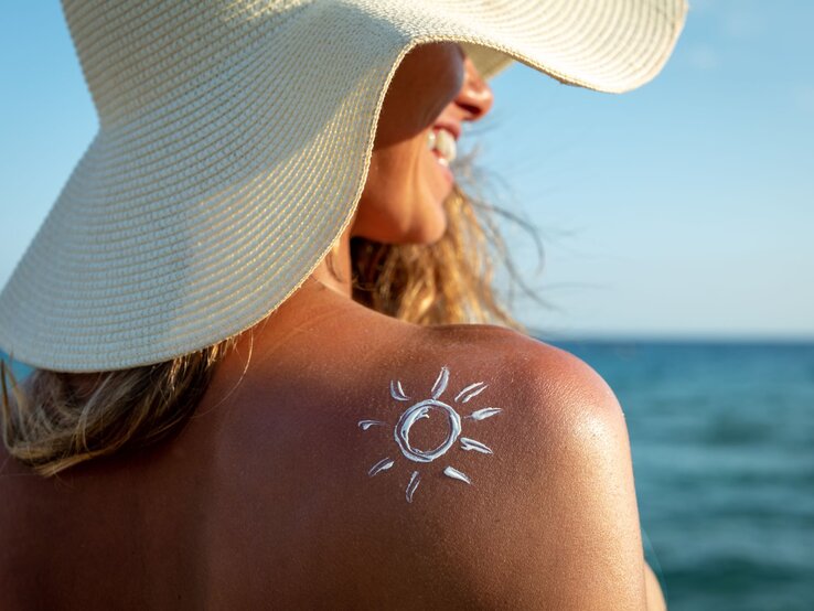 Junge Frau mit Strohhut und Sonnencreme-Symbol auf der Schulter lacht am Strand bei sonnigem Wetter.