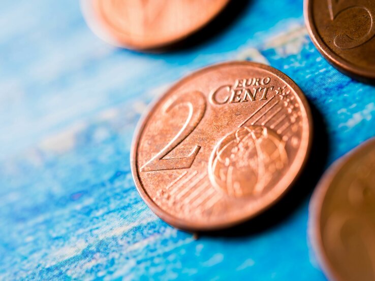 Eine Nahaufnahme einer 2-Cent-Euro-Münze auf einem blauen Untergrund, unscharf im Hintergrund weitere Münzen.