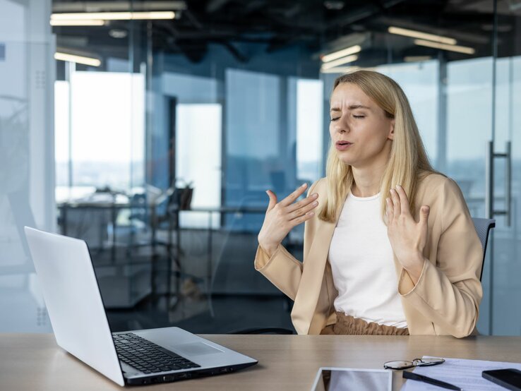 Eine blonde Frau in beigefarbener Bluse und weißem Shirt sitzt gestikulierend vor einem Laptop in einem modernen Büro.