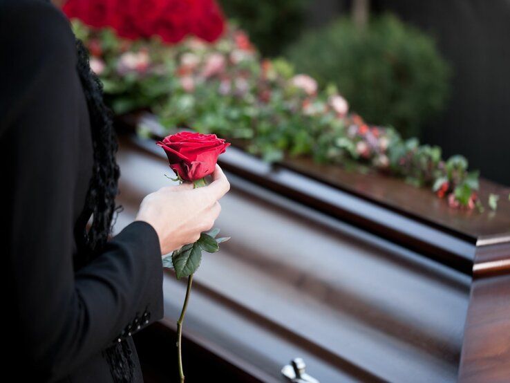 Eine Person in schwarzer Kleidung hält eine rote Rose vor einem mit Blumen geschmückten Sarg.