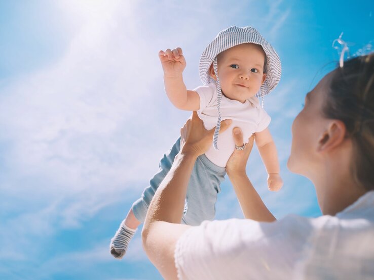 Junge Frau hebt lächelnd ein Baby mit gestreiftem Sonnenhut und weißem T-Shirt in den strahlend blauen Himmel.