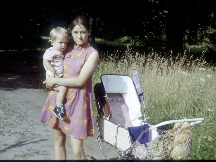 Mutter in lila Kleid mit Kind auf dem Arm, steht neben einem weißen Kinderwagen, im Hintergrund hohes Gras.