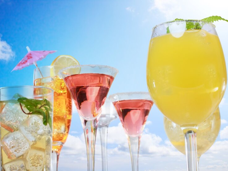 Bunte Cocktails mit Früchten, Minze und Eiswürfeln stehen vor strahlend blauem Himmel und weißen Wolken.