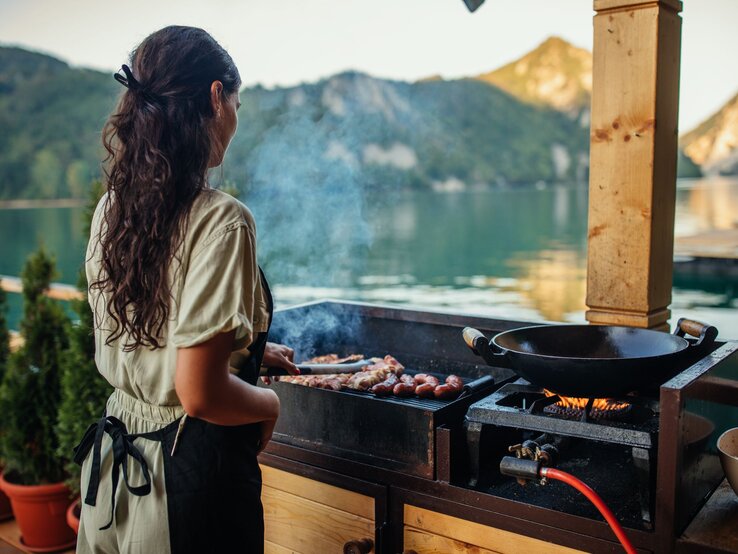 Eine junge Frau mit langen braunen Haaren grillt am Abend Würstchen an einem See mit Bergkulisse im Hintergrund.