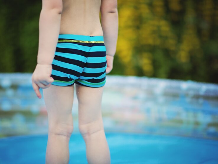 Ein Kleinkind in blau-schwarz gestreifter Badehose steht am Rand eines Planschbeckens mit verschwommenem grünen Hintergrund.