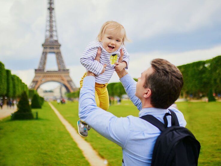 Ein lachender Vater hebt sein fröhliches Kleinkind vor dem Eiffelturm in Paris in die Luft, auf einer grünen Wiese.