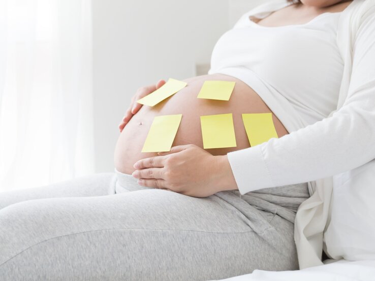 Schwangere Frau in grauer Hose und weißem Oberteil sitzt entspannt und hat gelbe Notizzettel auf ihrem Bauch.