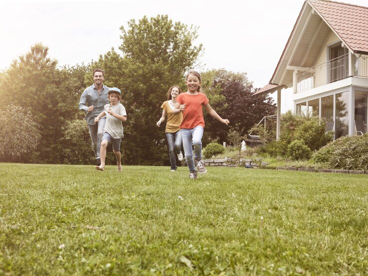 Eine Familie mit zwei Kindern rennt lachend über eine grüne Wiese vor einem modernen Haus an einem sonnigen Tag.