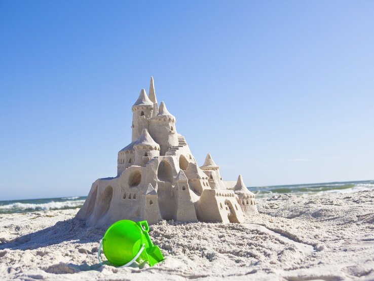 Große Sandburg mit vielen Türmen und Bögen steht am sonnigen Strand, ein grüner Eimer liegt im Vordergrund. | © Getty Images/Pam McLean