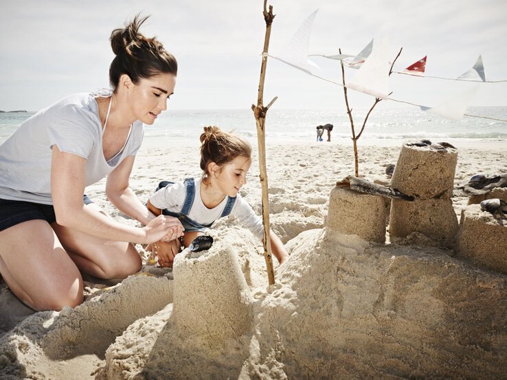 Eine junge Frau mit braunem Haar und ein Mädchen in Latzhose bauen konzentriert eine Sandburg am sonnigen Strand.