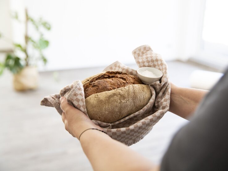 Zwei Hände halten ein frisch gebackenes Brot in einem beige karierten Tuch, daneben eine Schale mit Salz, vor unscharfem Hintergrund.