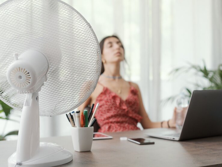 Junge Frau in rotem Sommerkleid sitzt erschöpft vor einem Laptop, während ein Ventilator auf sie gerichtet ist.