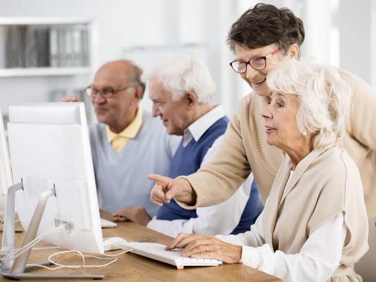 Eine ältere Frau zeigt einer anderen Frau etwas auf einem Computerbildschirm. Sie freuen sich. Zwei Männer im Hintergrund.