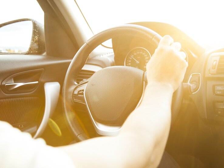 Eine Hand am Lenkrad eines Autos, in das warme, gleißende Sonnenlicht strahlt.