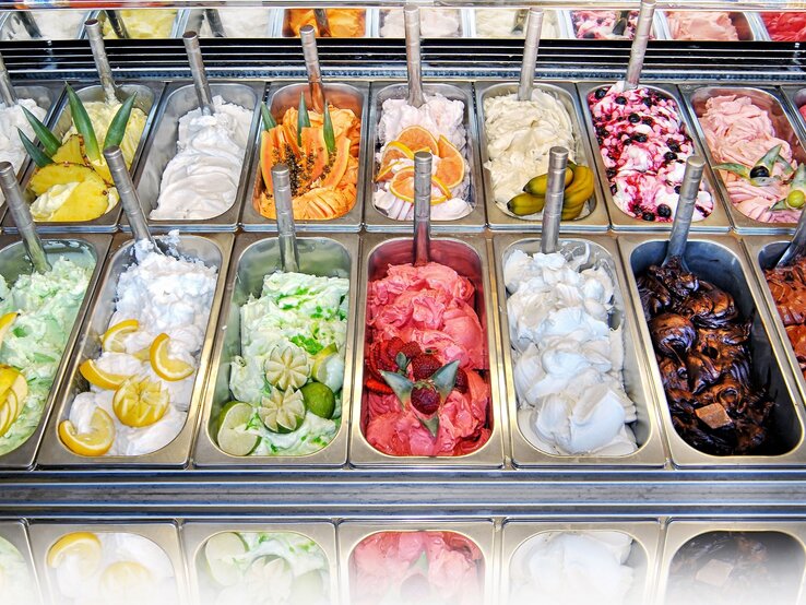 Verschiedene Sorten Eiscreme in Metallbehältern, dekoriert mit Obstscheiben, darunter Zitrone, Limette und Erdbeeren. | © iStock.com/Photology1971