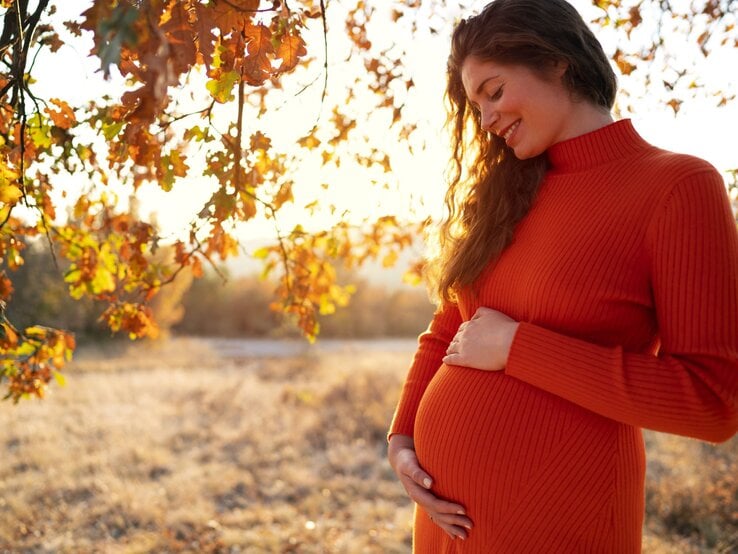 Junge schwangere Frau in einem orangefarbenen Kleid lächelt glücklich und hält ihren Bauch in einer herbstlichen Landschaft.