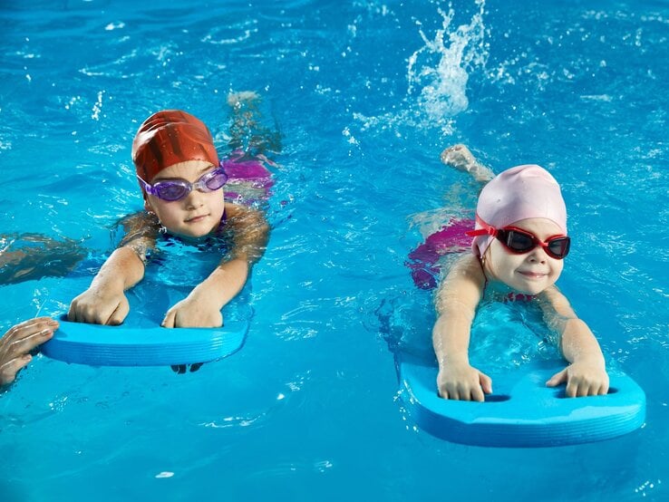 Zwei Kinder mit badekappe und schwimmbrille werden mit Schwimmbrettern an das Wasser gewöhnt.