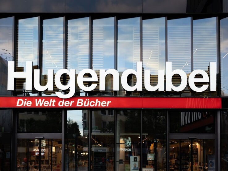 Große weiße Leuchtschrift "Hugendubel" mit rotem Slogan "Die Welt der Bücher" über der Glasfassade einer Buchhandlung.