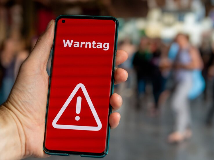 Hand hält Smartphone mit rotem Bildschirm und weißem Warnsymbol, darauf steht „Warntag“, im unscharfen Hintergrund Menschen.