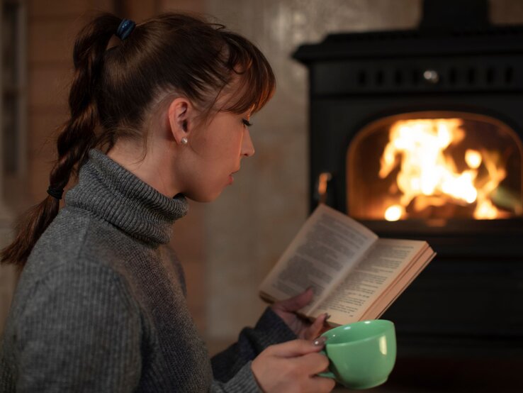 Junge Frau mit geflochtenem Zopf liest vertieft ein Buch vor einem Kaminfeuer und hält eine grüne Tasse in der Hand.