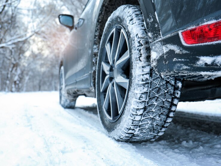 Ein dunkles Auto mit Schneereifen fährt auf einer verschneiten Straße durch einen winterlichen Wald mit kahlen Bäumen.