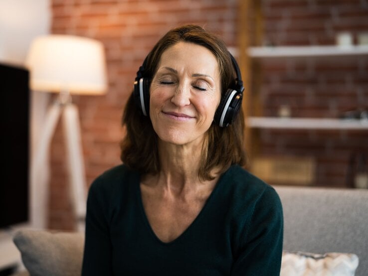 Eine lächelnde Frau mittleren Alters mit geschlossenen Augen genießt Musik über große Kopfhörer in einem gemütlichen Wohnzimmer.