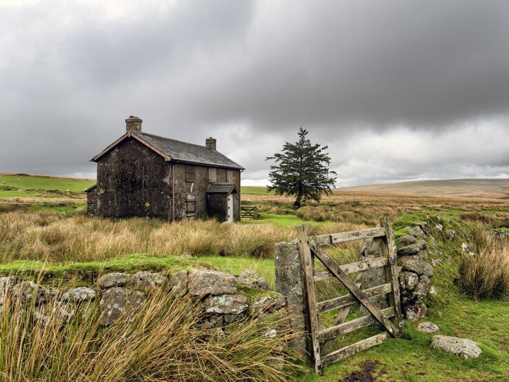 Ein verlassenes Steinhaus steht unter düsteren Wolken in einer abgelegenen, windgepeitschten Moorlandschaft neben einem Holzzaun.