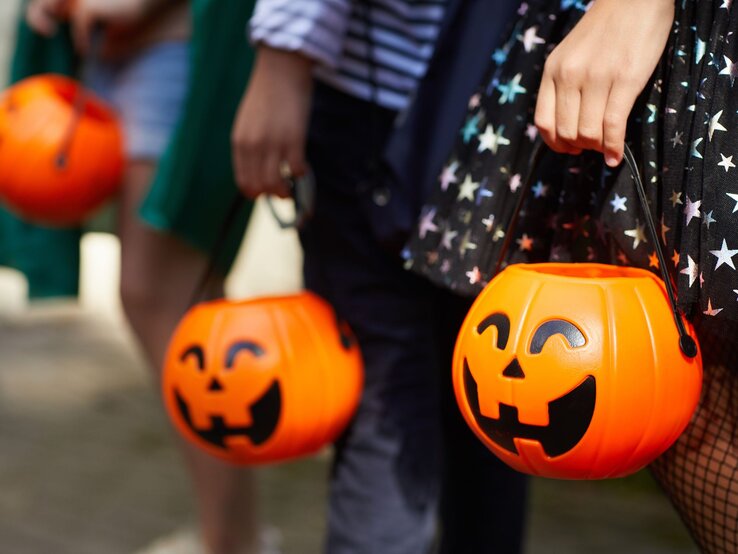 Kinder halten orangefarbene Halloween-Kürbiseimer in der Hand, während sie in bunt gemusterten Kostümen auf Süßigkeitensuche gehen. | © AdobeStock/AnnaStills