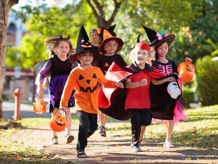 Eine Gruppe verkleideter Kinder läuft lachend auf einem Gehweg, alle halten Halloween-Körbchen in den Händen. | © AdobeStock/famveldman