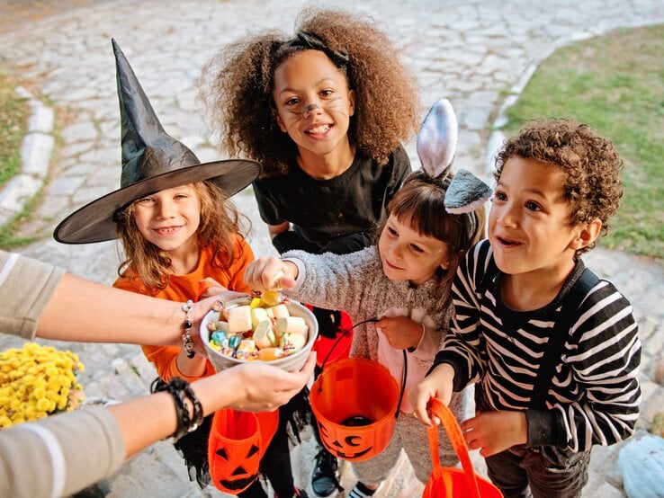 Vier fröhliche Kinder in Halloween-Kostümen halten orangefarbene Eimer und nehmen Süßigkeiten aus einer Schale entgegen.